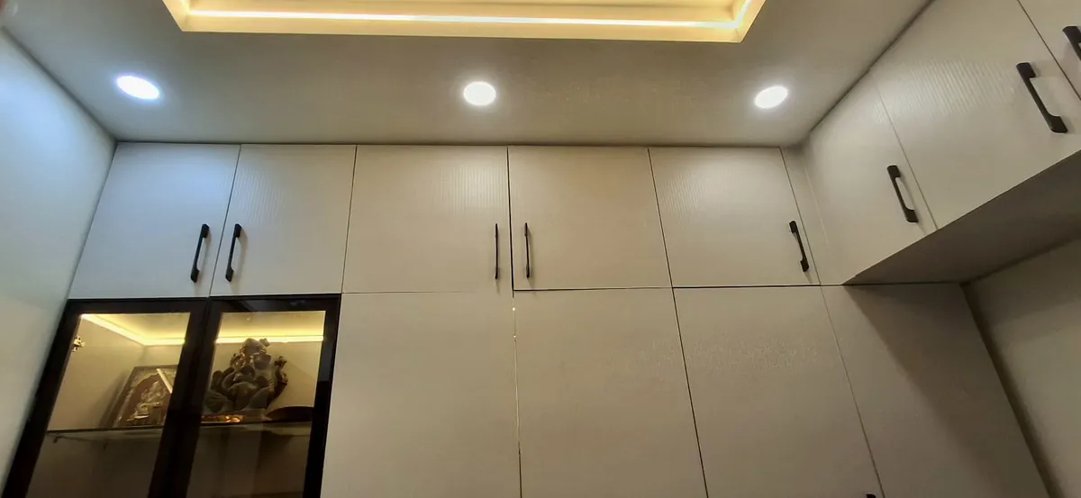 Built-in pooja niche with a lit glass-front display housing a Ganesha idol, set within a run of cream wardrobe cabinetry.