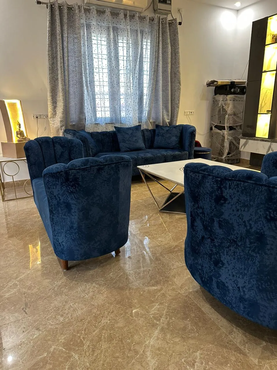 Living room part-way through handover, with a blue crushed-velvet sofa and two tub chairs round a marble-top table on a chrome X-frame