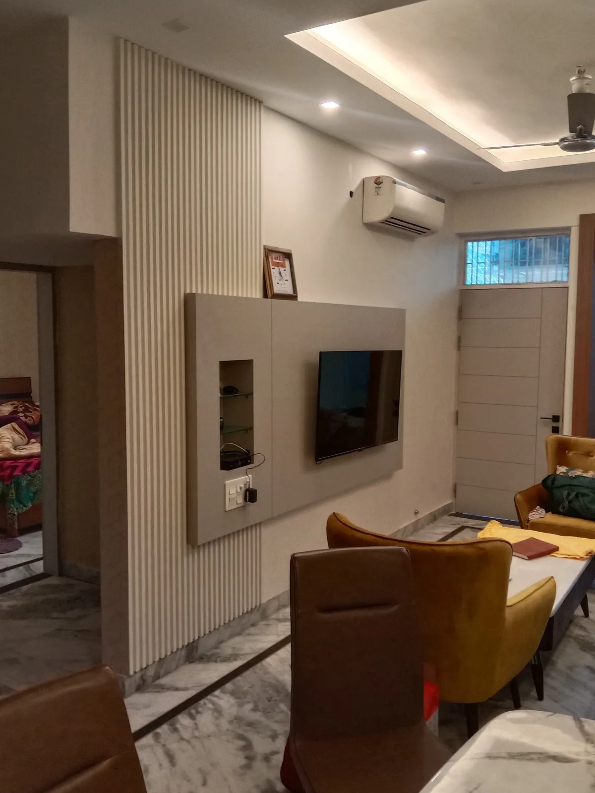 Dining corner facing a beige TV panel with a lit glass-shelf niche, set against floor-to-ceiling white fluted panelling