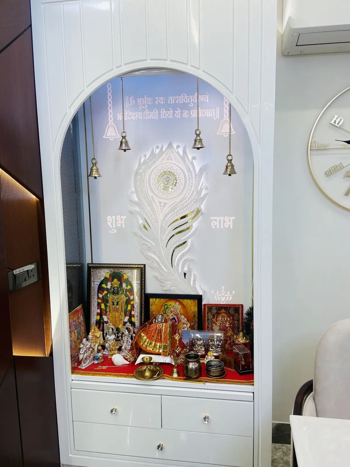 Built-in white lacquered pooja unit with an arched backlit niche, a peacock-feather motif panel, hanging bells, and idols set on a red cloth.