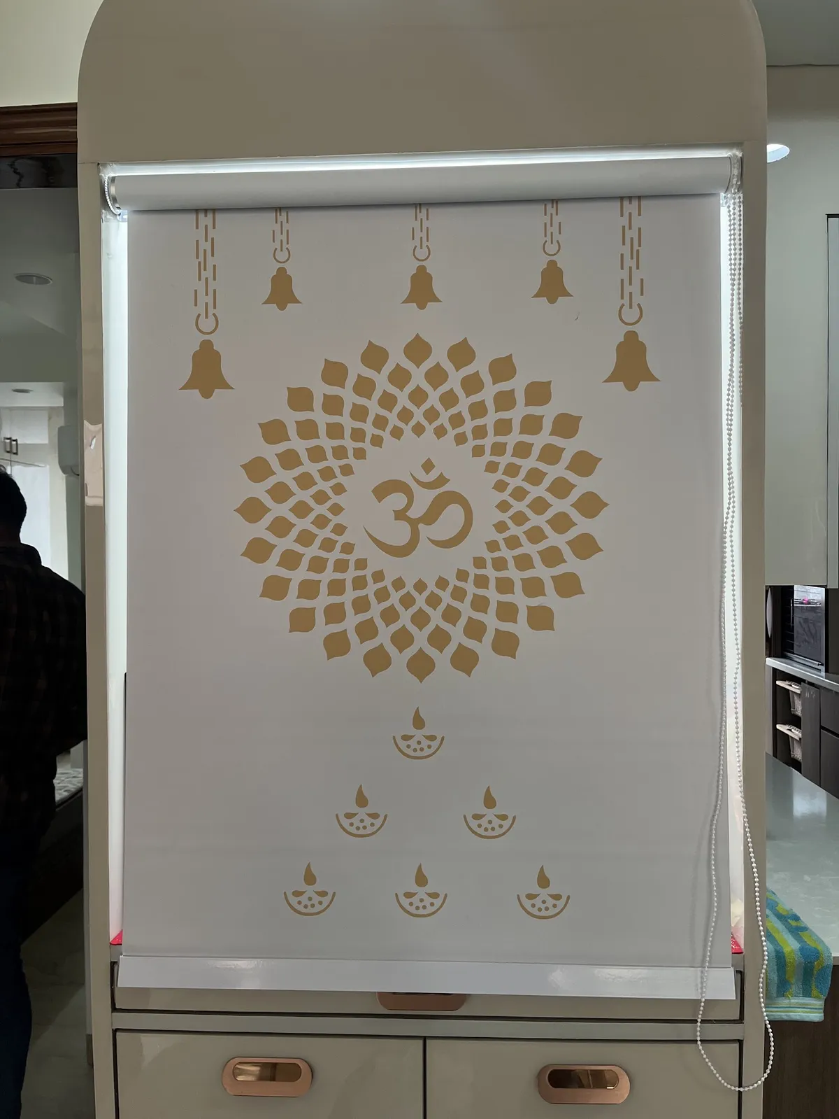 White pooja cabinet fitted with a printed roller-blind shutter featuring an Om motif, bells, and diya patterns, with storage drawers below.