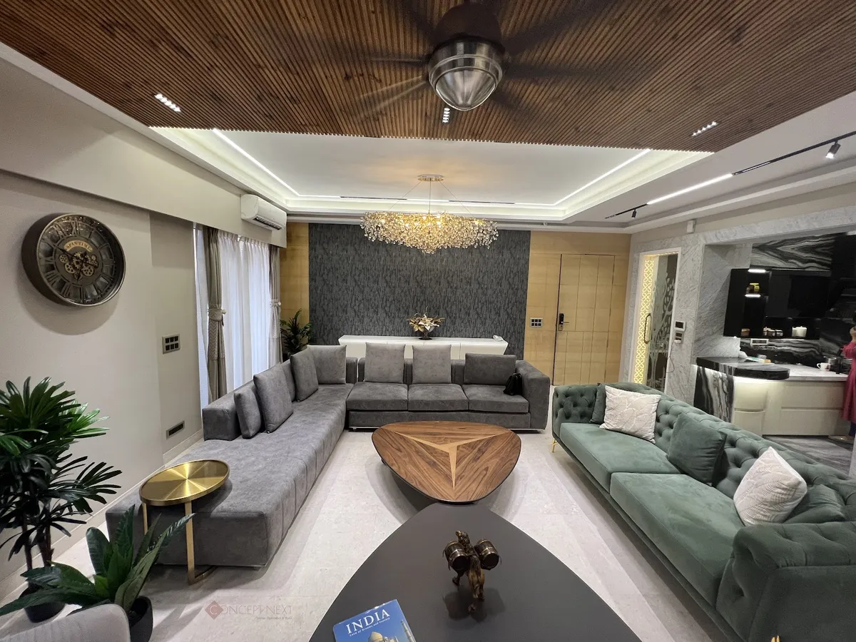 Living room with an L-shaped grey sofa facing a tufted sage-green sofa, a triangular wood coffee table, a crystal chandelier under a wood-slat ceiling