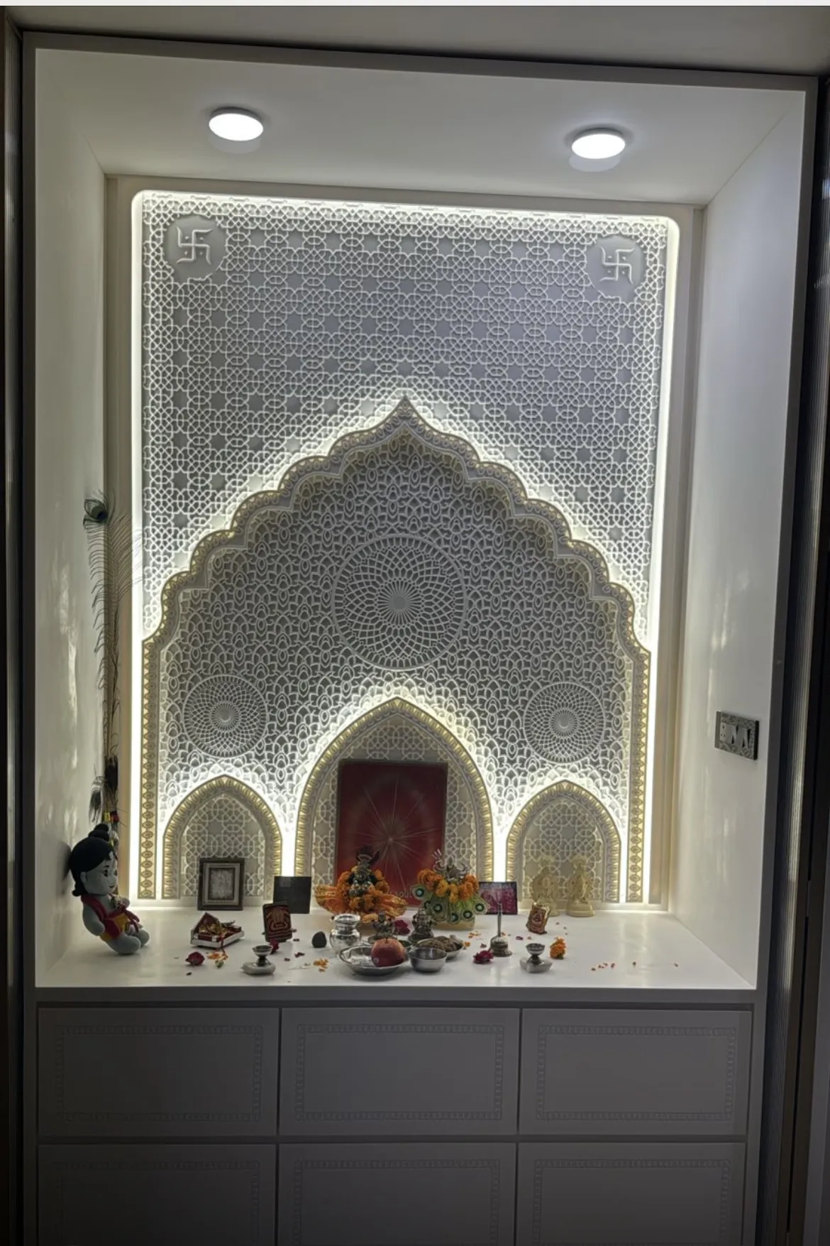 Backlit white CNC lattice pooja niche with a cusped Mughal arch and swastik corner medallions
