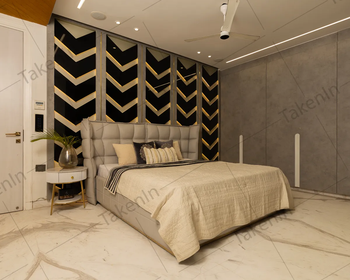 Master bedroom with a backlit black-and-gold chevron panel wall behind a tall grey biscuit-tufted headboard, a fluted round bedside on brass legs