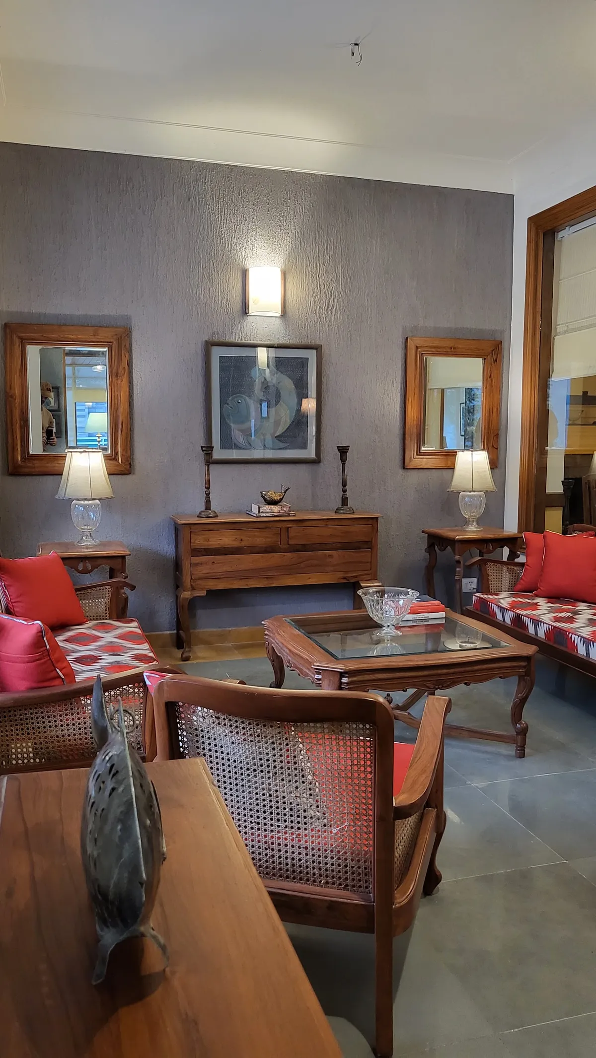 Sitting room against a coarse grey textured wall, a pair of teak-framed mirrors flanking a framed fish painting over a teak chest