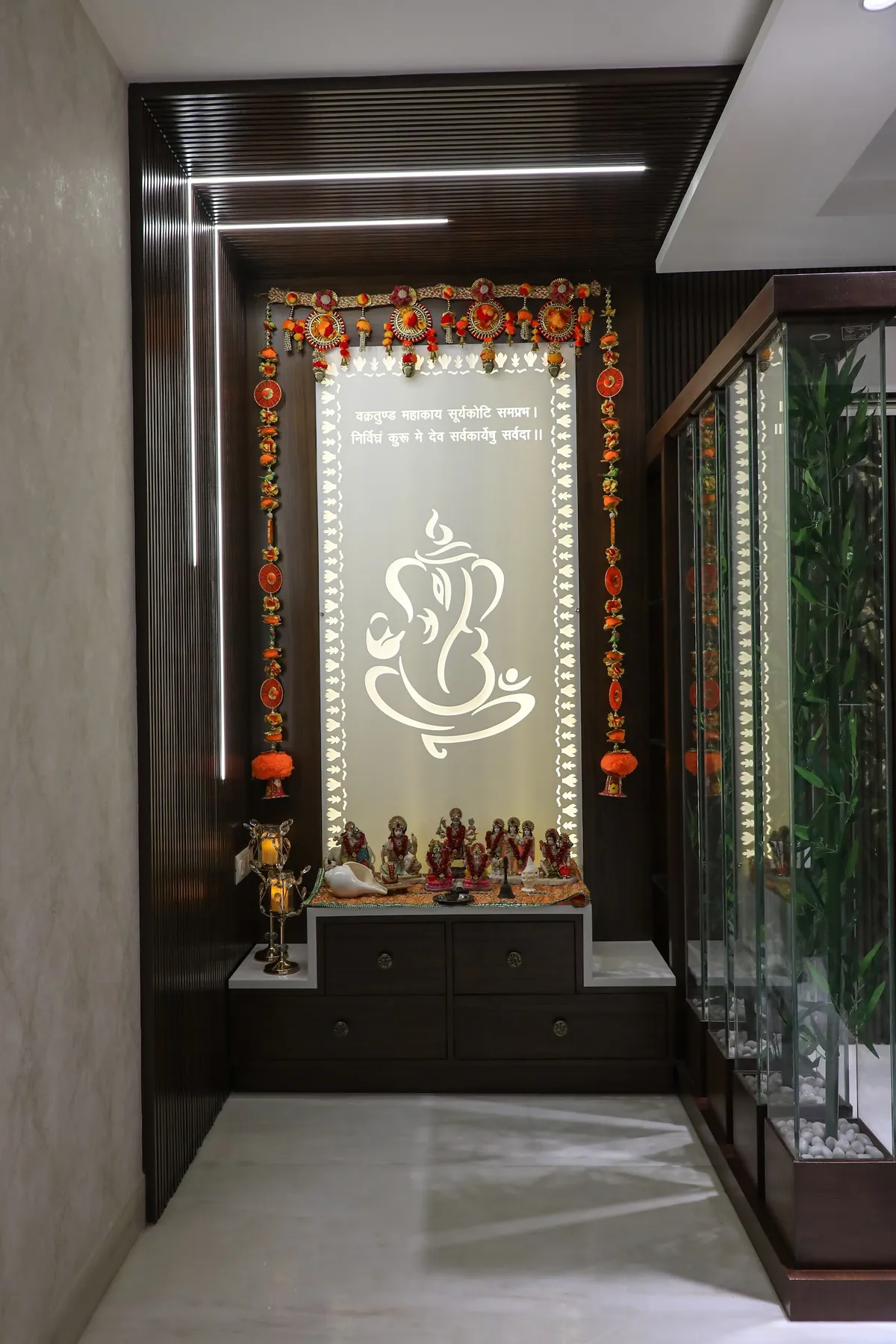 Pooja alcove framed in dark fluted wood with recessed linear lights running the frame: a backlit panel of hanging-bell cutwork and a line-art Ganesh