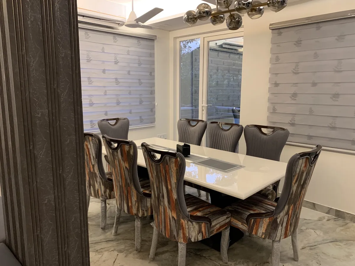 An eight-seater dining room with a white glossy table, patterned wingback dining chairs, a chrome sputnik chandelier and a marble-clad column.