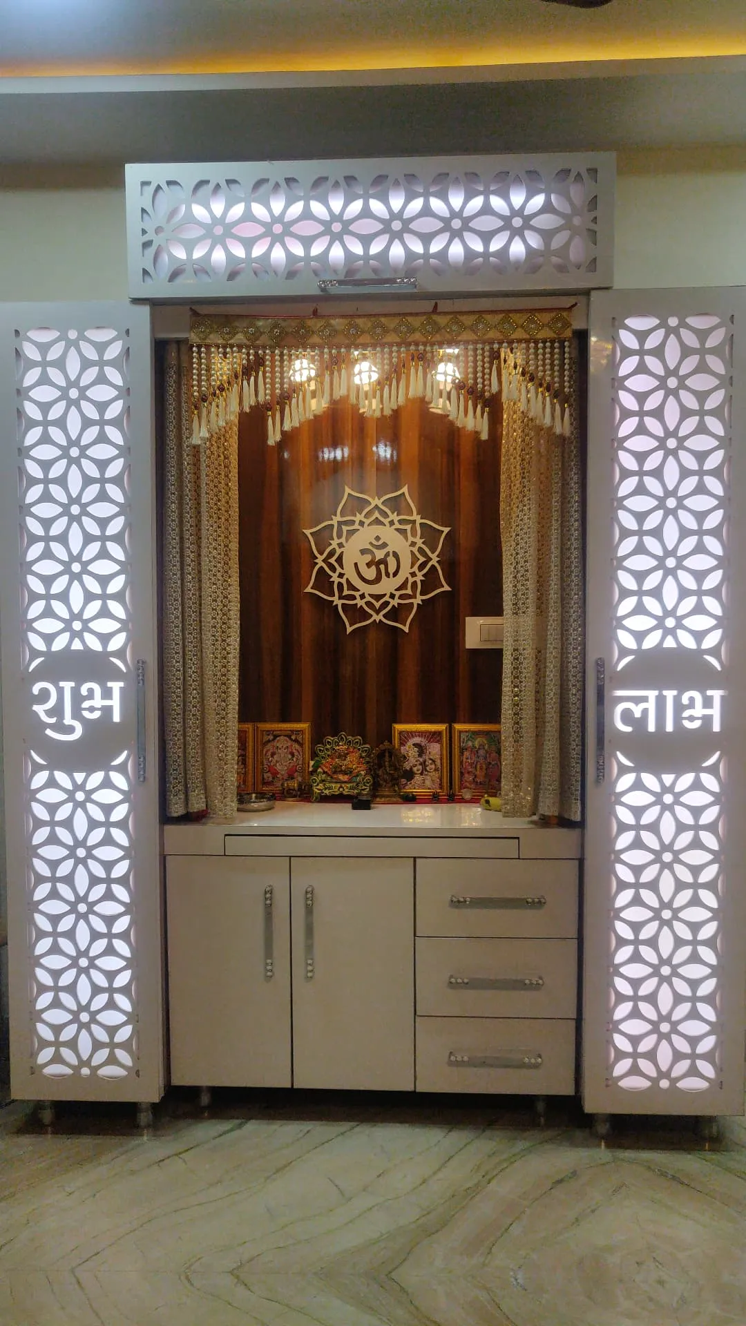 Dedicated pooja unit with backlit floral jaali side panels, an Om medallion backdrop, and cabinet storage below with deity idols on the ledge.