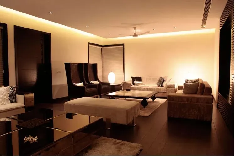 A dimly lit living room with dark wood flooring, tall wingback accent chairs, and a beige sectional sofa around a glossy black coffee table