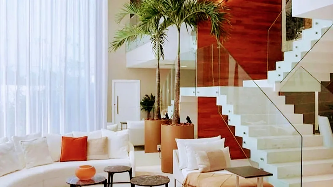 Double-height living room with a curved white sofa and orange accent pillow, tall potted palms flanking a floating staircase with glass balustrade and a rich