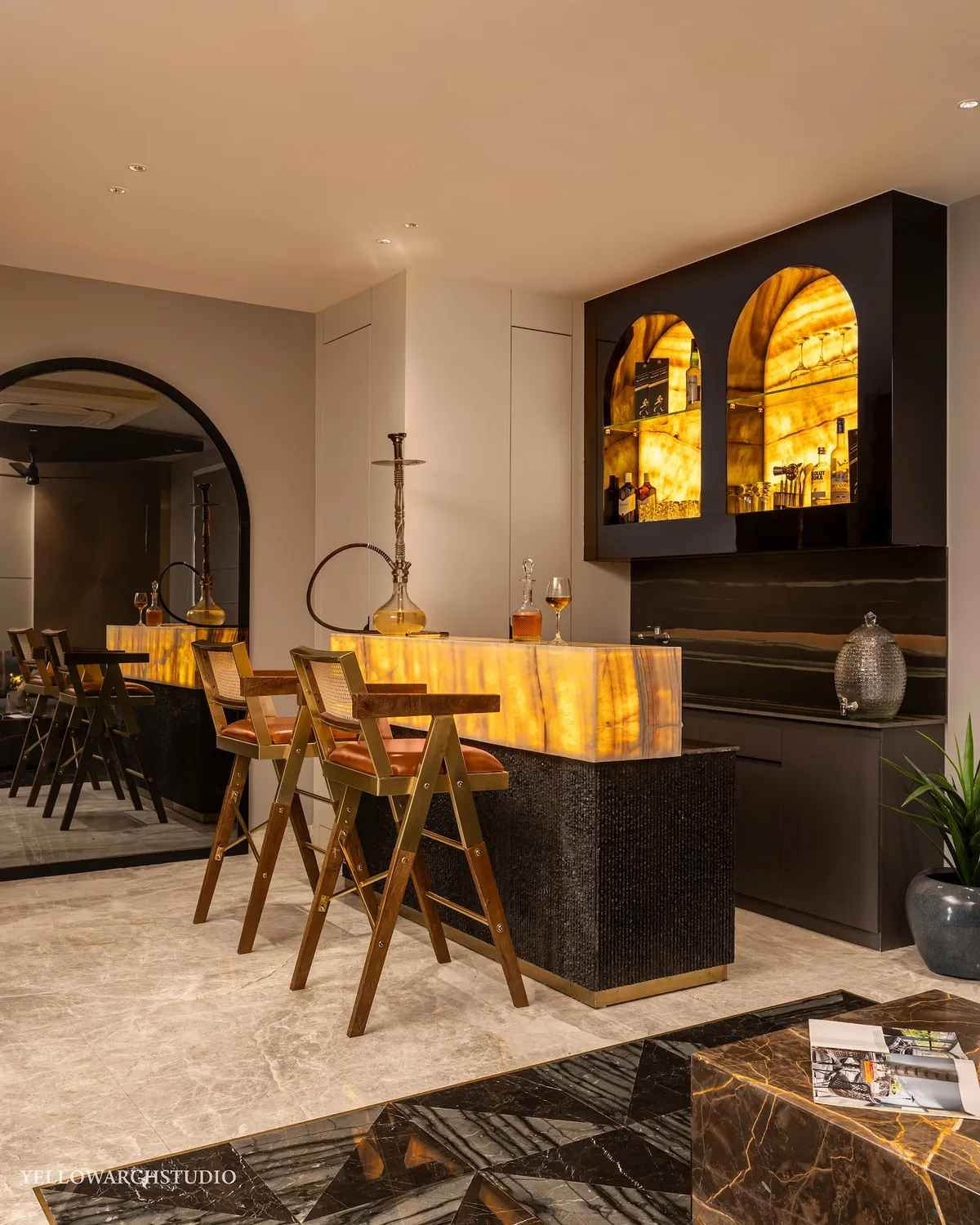 Home bar with a backlit honey-onyx counter on a fluted black base with brass toe-kick, twin arched onyx-lit liquor niches
