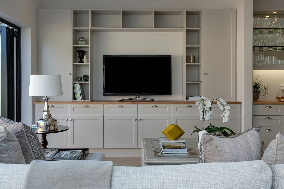 Living room built-in TV wall with shaker-style cabinetry, open display niches and a wood counter, styled sofa and glass coffee table in the foreground.