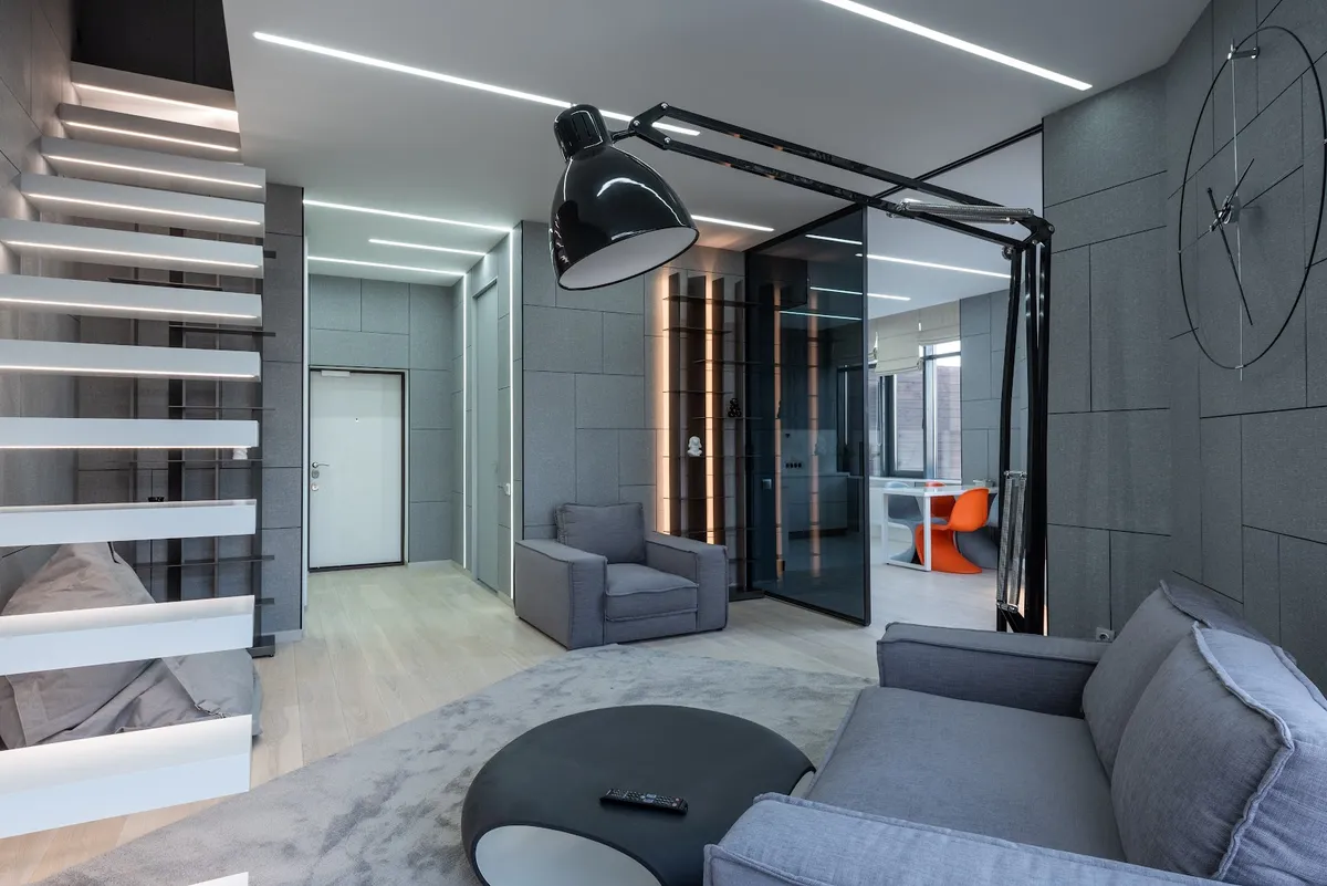 A grey upholstered-wall living room with an oversized articulated black task lamp over the sofa and a glass partition leading to a dining nook with orange