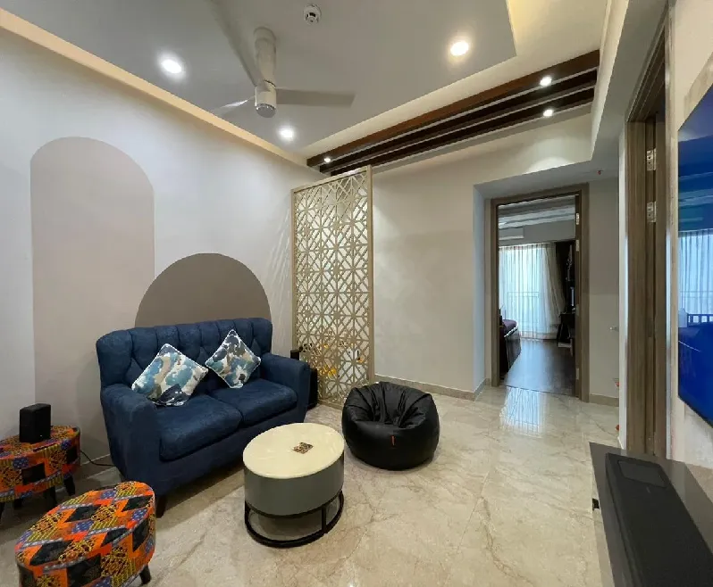 Living room with a navy tufted loveseat, colourful patterned ottomans, a black bean bag, and a gold-toned decorative jaali partition beside a painted wall arch
