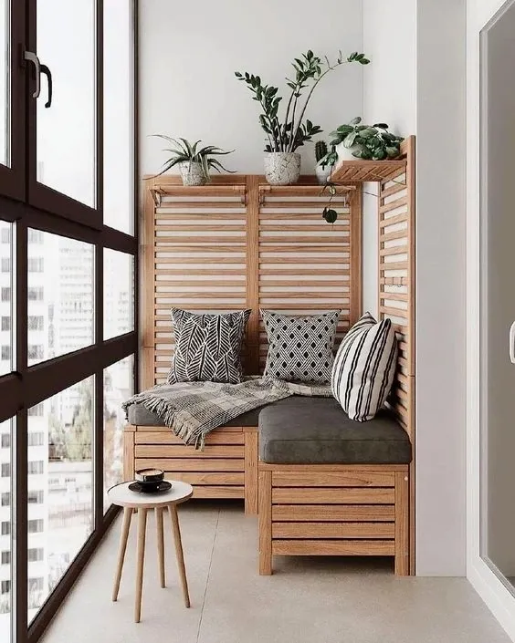 Corner of a glazed high-rise balcony with an L-shaped slatted-wood storage bench, black-and-white patterned cushions and potted greens lined along its shelf