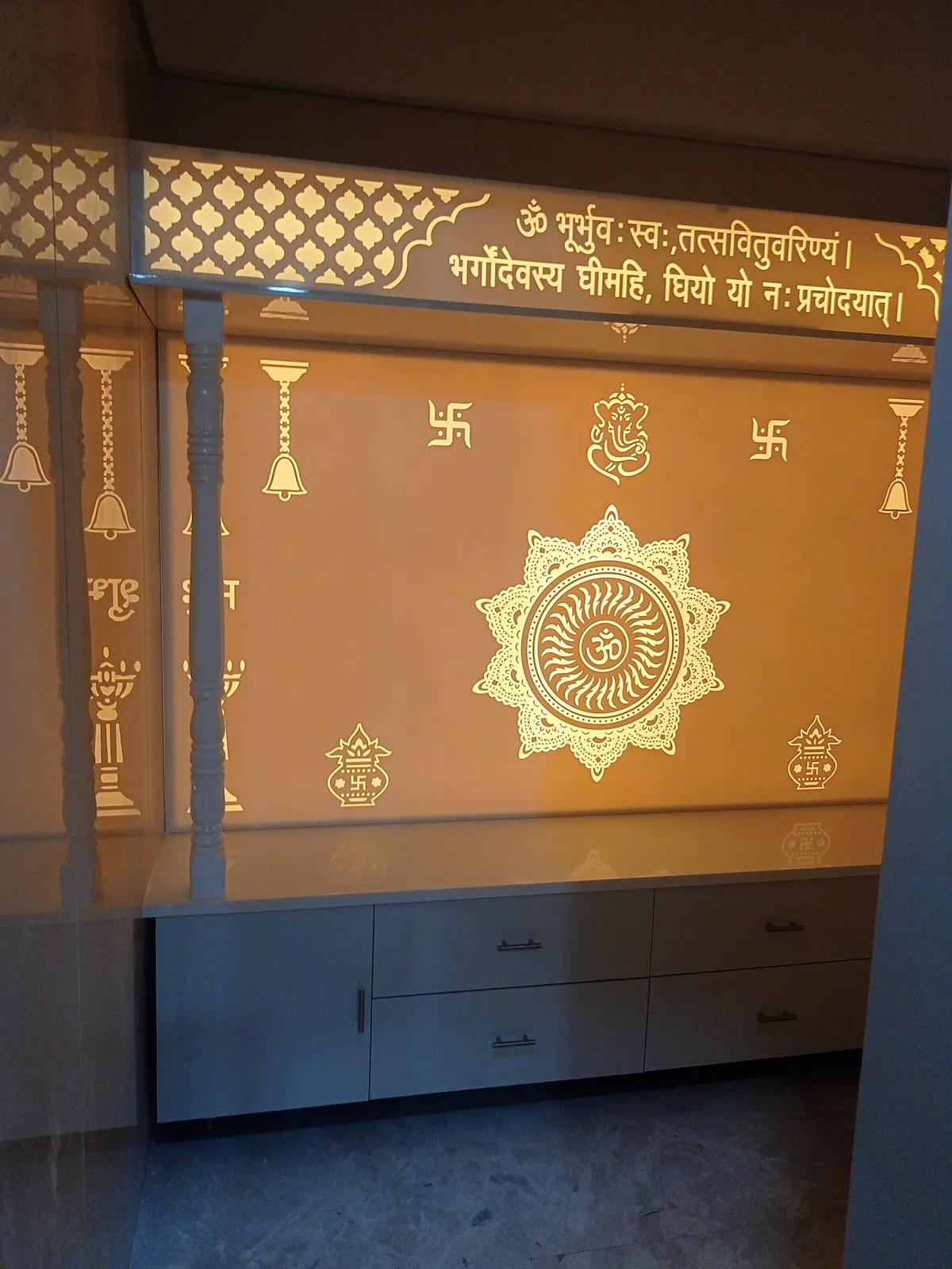 A pooja room with a backlit amber glass panel etched with the Gayatri mantra, an Om mandala and Ganesh motifs above a white storage cabinet