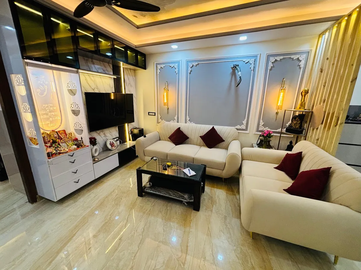 Living room whose end wall combines a backlit white jaali pooja unit with an Om cut-out and deity idols, a TV on marble-look cladding and glass-fronted lofts