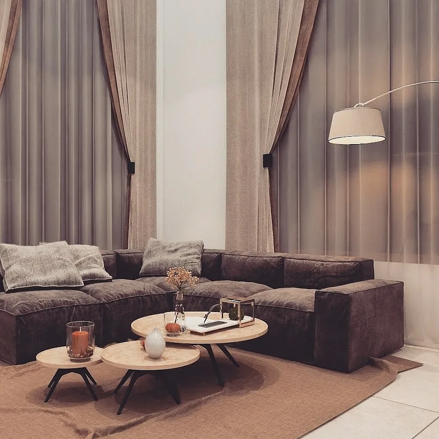 Living room with a dark brown modular sofa, floor-to-ceiling taupe curtains, a brass arc floor lamp, and nested round coffee tables on a jute rug.