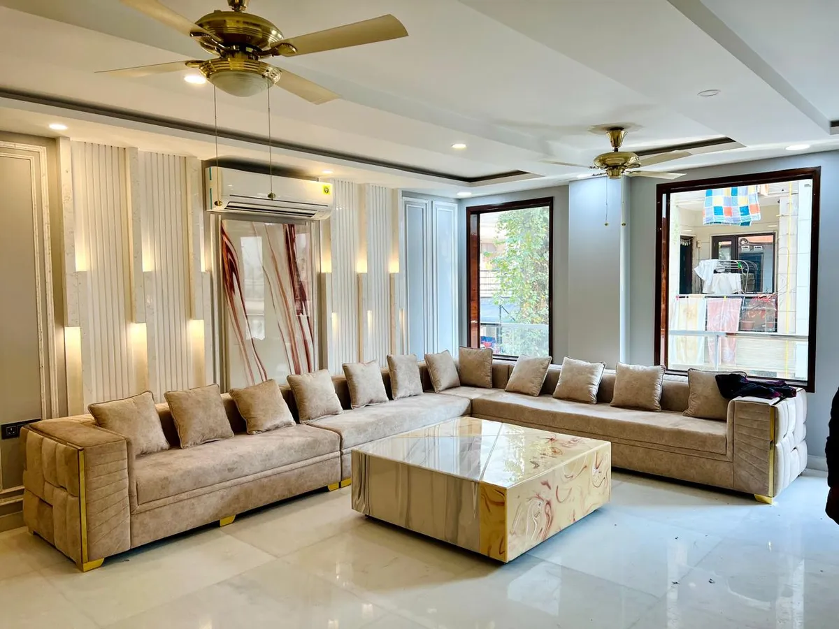 Long beige velvet L-sofa with gold-trimmed bases facing a red-veined onyx-look block coffee table