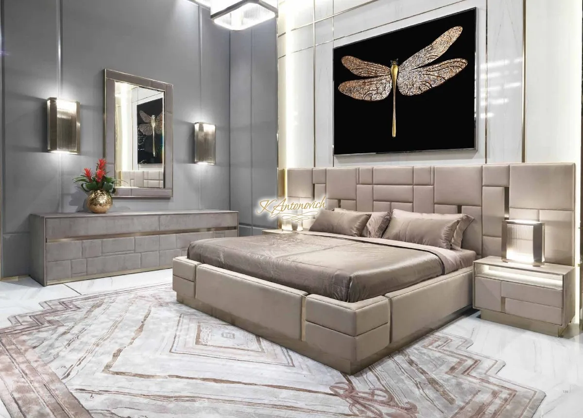 Render of a bedroom with a tufted taupe upholstered bed and panel headboard, a dragonfly art piece, and a mirrored dresser with brass trim.