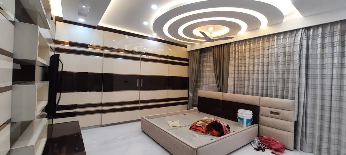 Bedroom photographed at handover, bare bed base still holding paint buckets and cloth, under concentric circular POP rings that ring the ceiling fan
