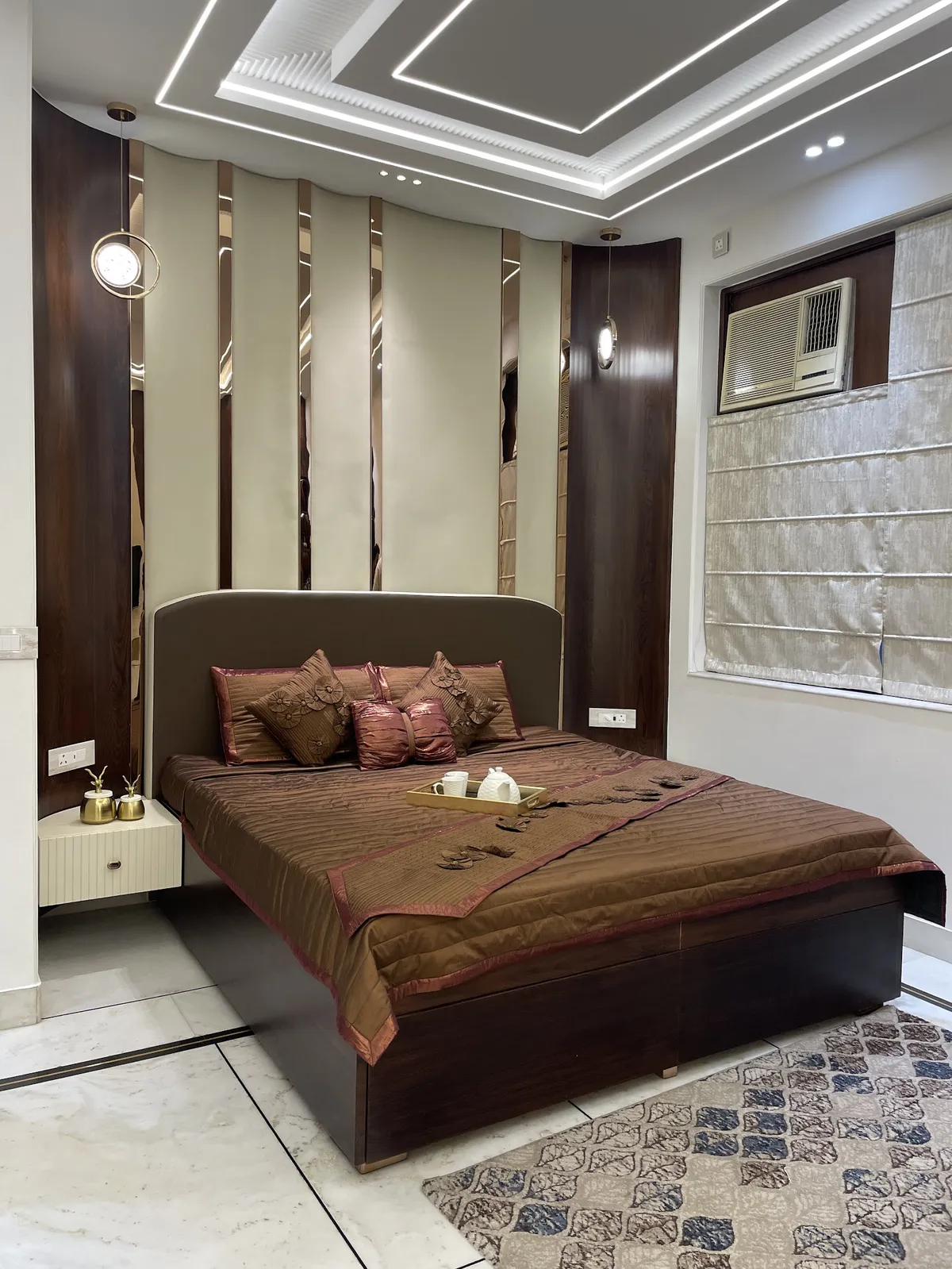 A luxury bedroom with a dark wood and mirrored-strip panelled headboard wall, a geometric LED cove ceiling, bronze bedding with a styled welcome tray