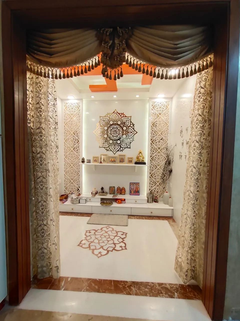 Walk-in pooja room with a gold laser-cut mandala above a backlit white niche, jaali side panels, a tasselled swag valance across the walnut-framed opening