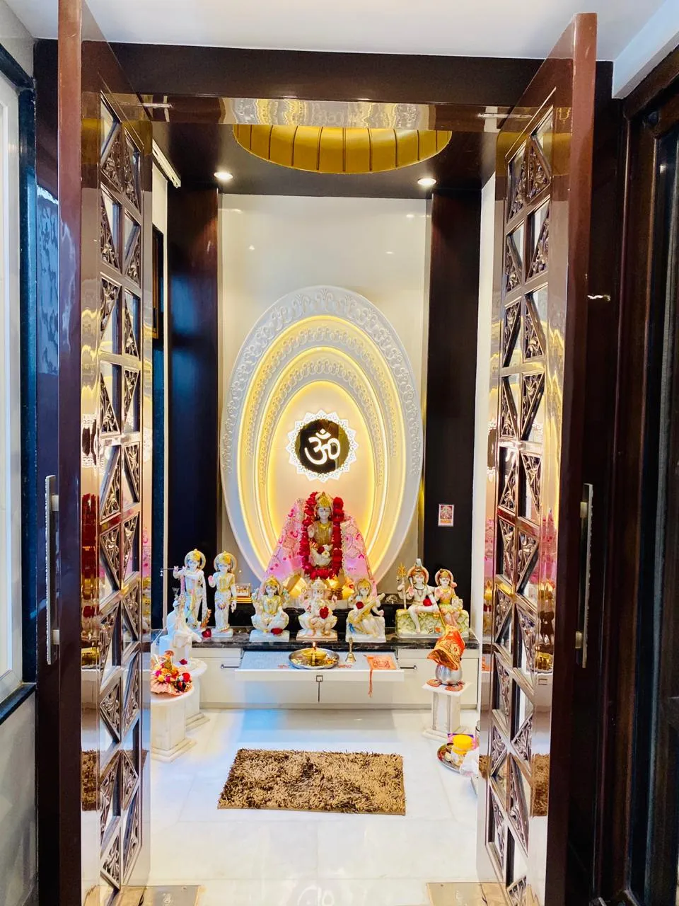 Dedicated pooja room seen through mirror-inlay lattice double doors, with idols on a marble platform against a concentric carved oval backdrop lit warm behind