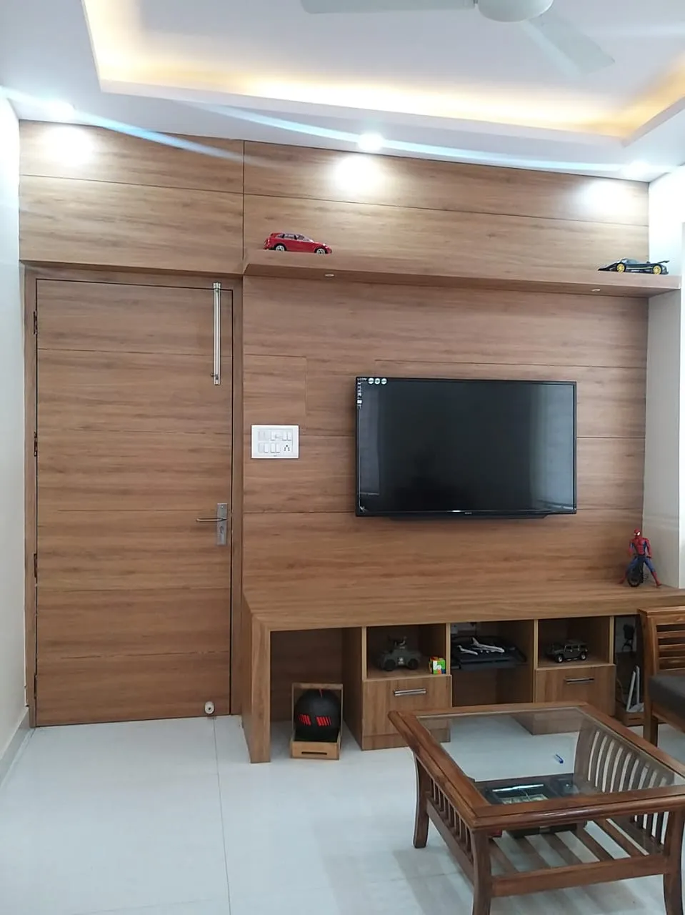 Living room where a teak-laminate wall runs unbroken from the flush door across the TV panel