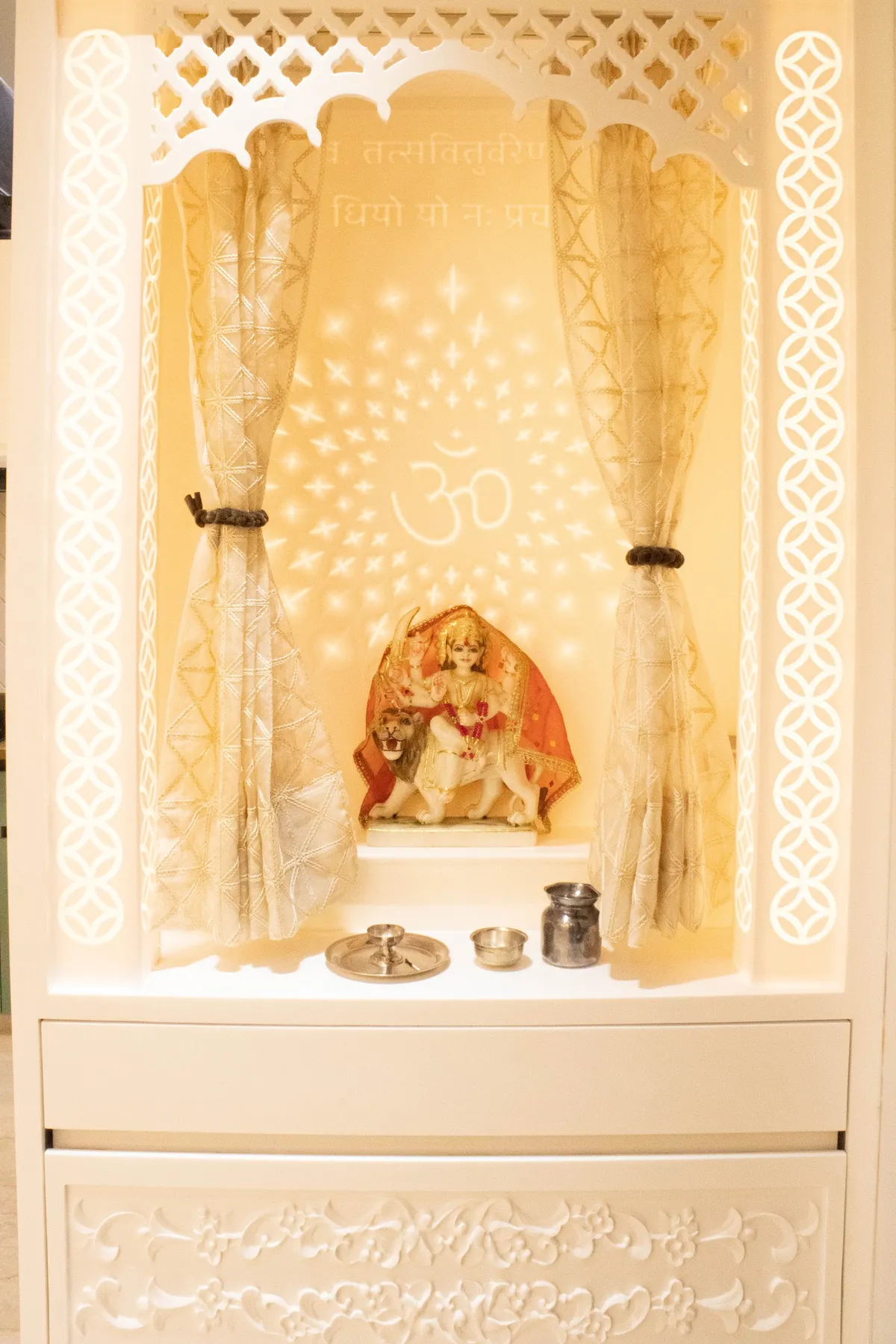 Close-up of a white pooja unit niche with a carved jaali arch, sheer gold tie-back curtains, and a backlit star-and-Om projection behind a Durga idol.
