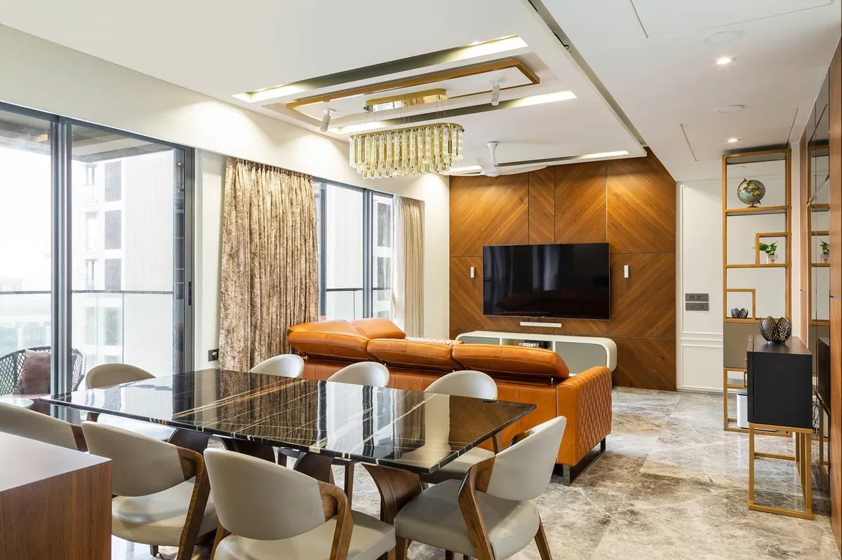 Open living-dining in a high-rise flat: a chevron walnut TV wall facing a tan quilted-leather sofa, a black marble dining table for eight in the foreground