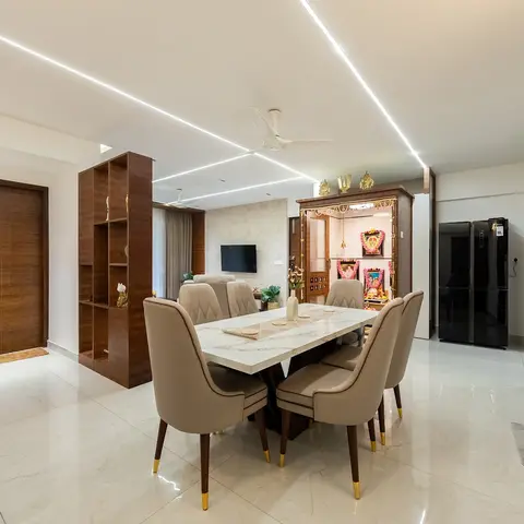 An open-plan home showing a marble dining table, a wood-framed pooja unit, and a living room with sofa and TV, linked by a