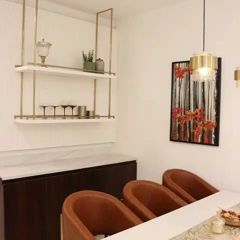 Dining nook with tan leather barrel chairs around a white marble table, a brass-framed open bar shelf styled with glassware,
