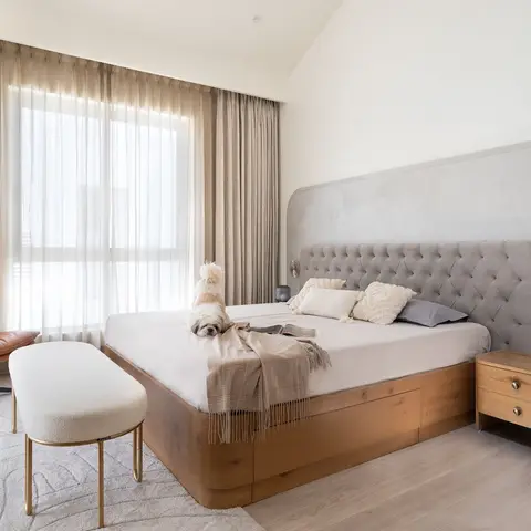 a small dog sits on a king bed with a tufted grey headboard against a curved plaster wall, tan leather swivel chair in the