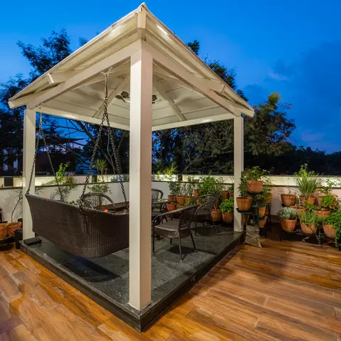 A rooftop terrace at dusk with a white timber gazebo sheltering a hanging rattan swing bed and a rattan dining set,
