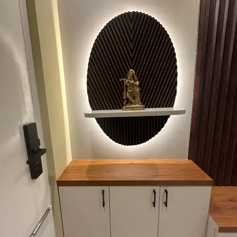 Foyer wall niche with a backlit oval fluted-wood panel framing a brass Radha-Krishna idol on a floating shelf, above a