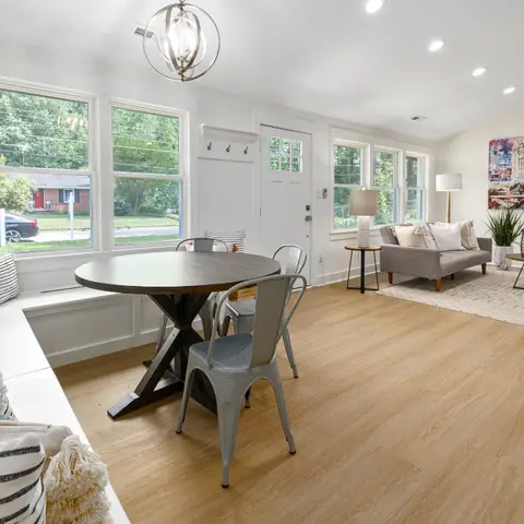 Open-plan dining and living area with a round wood trestle table and metal chairs under an orb chandelier, a built-in window