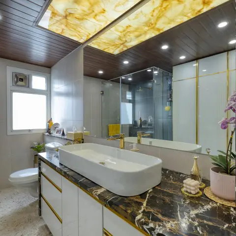 A luxury bathroom with a backlit amber onyx ceiling panel set into a dark wood slat ceiling, a black-and-gold marble vanity