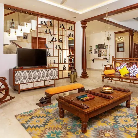 a double-height Indian living room with a carved wooden swing, rocking chair, and a low carved coffee table with brass diyas