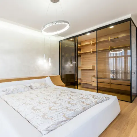 Bedroom with a curved white platform bed beneath a double-ring LED pendant chandelier, beside a black-framed glass walk-in