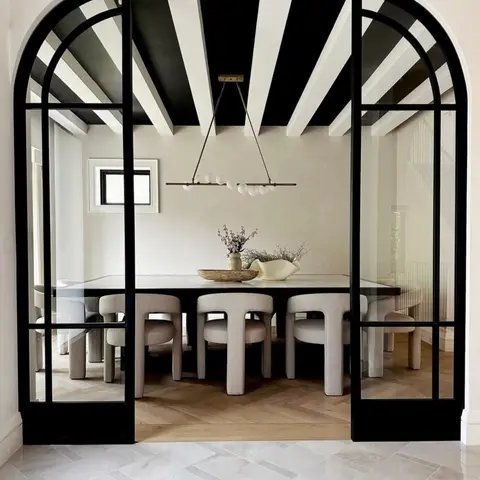 Dining room seen through arched black-framed French doors, with a black-and-white painted exposed-rafter ceiling and a