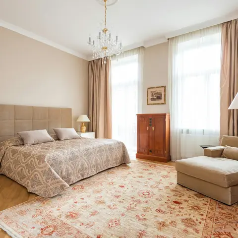 a grand master bedroom with a crystal chandelier, floor-length silk drapes, an upholstered headboard, a chaise lounge, and a