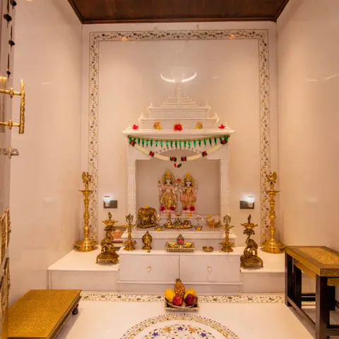 White marble pooja room with a carved temple-style mandir alcove housing deity idols, brass diyas and elephant statues