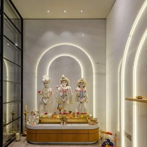 Pooja room with a backlit marble arch niche housing deity idols, a fluted wood cabinet base, brass diyas, and a glass