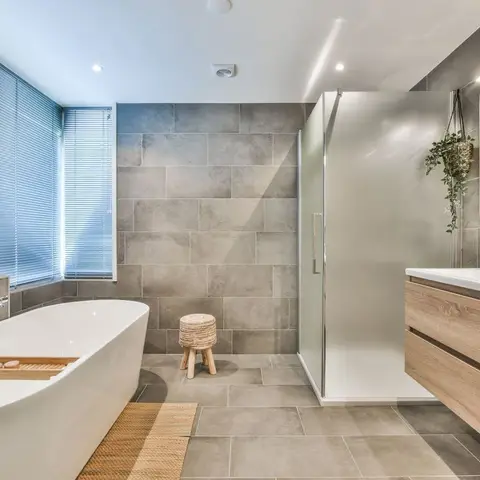 A bathroom with a freestanding oval tub beneath louvre blinds, large-format grey stone-look tile walls, a frameless glass
