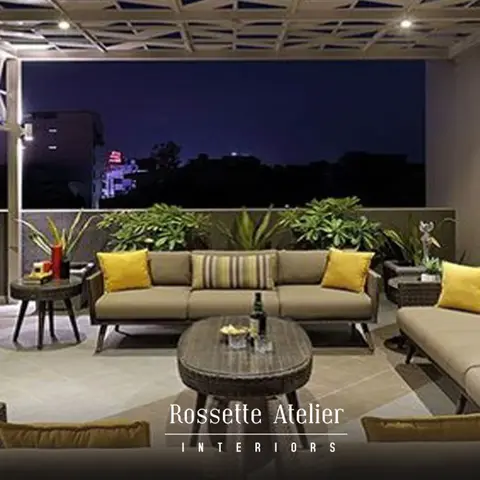 A rooftop terrace lounge with wicker sofas and mustard-yellow cushions beneath a geometric lattice pergola, potted plants