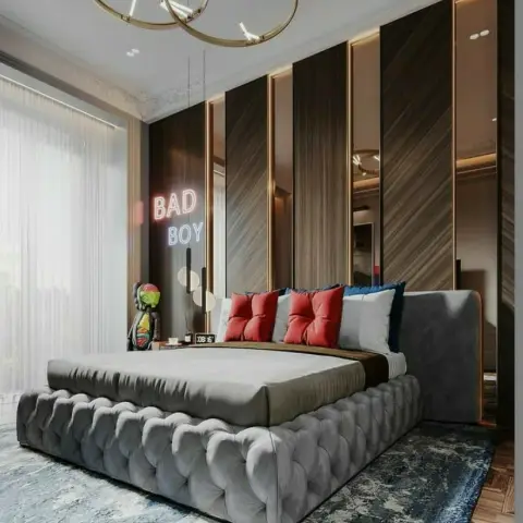 Bold bedroom with a grey tufted velvet bed, red and blue jewel-tone cushions, fluted dark-wood headboard wall with brass