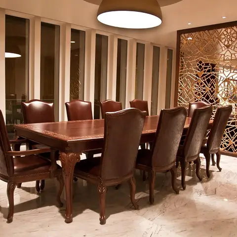 Ten-seat carved wood dining table with burgundy leather chairs on a marble floor, beside an ornate gold jaali partition