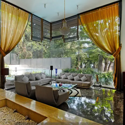 A double-height villa living room with floor-to-ceiling glazing onto a private garden and pool, gold silk drapery, a crystal