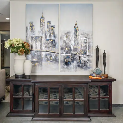 A dark wood console table styled with ceramic vases and bronze figurines beneath a large two-panel cityscape painting, set